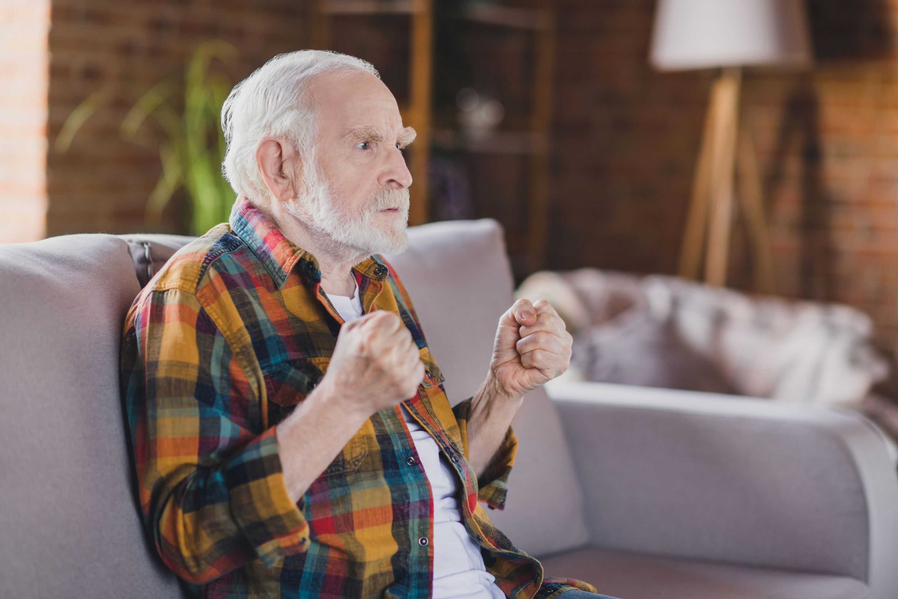 Senior bearded man sits looking angry with balled fists raised.
